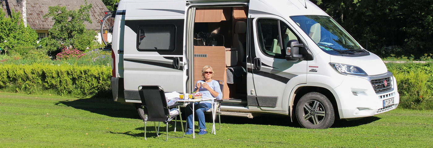Frau sitzt an einem Campingtisch mit Kaffee und Kuchen vor einem Wohnmobil auf einer Wiese